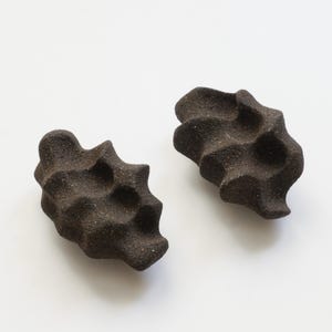 Può includere: Due oggetti in ceramica marrone scuro, strutturati e di forma organica. Ognuno ha una forma irregolare unica e una superficie ruvida e macchiettata. Sono posti su uno sfondo bianco liscio, evidenziando le loro qualità scultoree.
