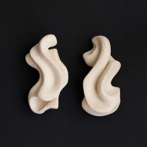 Könnte beinhalten: Zwei cremefarbene, abstrakte Skulpturen mit strukturierten Oberflächen vor schwarzem Hintergrund. Die Formen haben ein fließendes, organisches Design, das stilisierten Wellen oder Wolken ähnelt. Die Formen sind nebeneinander positioniert.