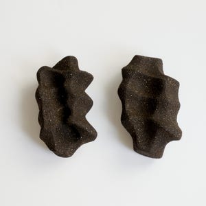 May include: Two dark brown, textured ceramic sculptures with irregular, organic shapes. The pieces have a rough, speckled appearance and are set against a plain white background. The forms have a unique, abstract design.