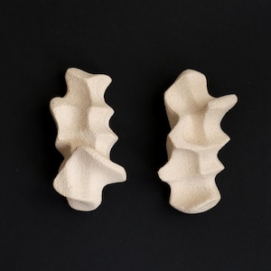Può includere: Due sculture astratte bianco sporco con superfici strutturate sono esposte su uno sfondo nero. Le forme hanno un aspetto stratificato e organico, che suggerisce un'estetica naturale o artigianale. Le sculture sono posizionate fianco a fianco.