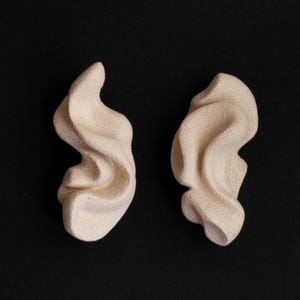 Può includere: Due forme scultoree astratte bianco sporco su sfondo nero. Le forme hanno un design fluido e organico, con una superficie strutturata. Le forme sembrano essere realizzate in un materiale solido, possibilmente ceramica o gesso.