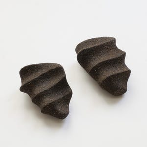 Può includere: Due pezzi in ceramica marrone scuro, strutturati, con una forma curva unica e un design a coste. Gli oggetti sono posti su uno sfondo bianco semplice, evidenziando la loro forma e consistenza. I pezzi sembrano fatti a mano.