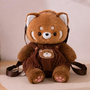 以下が含まれることがあります： 茶色のレッサーパンダのぬいぐるみバックパック。コーデュロイのオーバーオールデザインとクマのパッチが付いています。バックパックには調節可能なストラップと笑顔の顔があります。かぎ針編みのヒマワリも近くにあります。