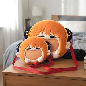 以下が含まれることがあります： オレンジ色の漫画のキャラクターの形をした2つのぬいぐるみバッグ。黒いアクセントと赤いストラップが付いています。バッグは口が開いており、木製の表面に置かれています。肩にかけるように設計されています。