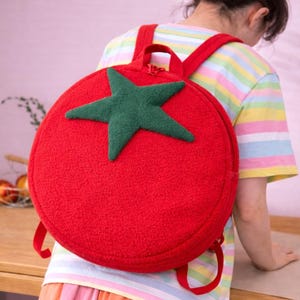 Puede incluir: Una mochila redonda roja con una estrella verde. La mochila tiene correas rojas y un asa superior. La bolsa está hecha de un material texturizado, que se asemeja a un tomate. La mochila está diseñada para niños.
