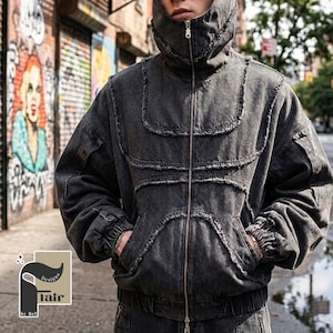 Könnte beinhalten: Schwarze Jeansjacke mit Reißverschluss und Kapuze. Die Jacke hat abgenutzte Details und Taschen. Die Jacke ist Teil eines passenden Sets. Der Hintergrund zeigt Graffiti-Kunst auf einer Backsteinmauer.