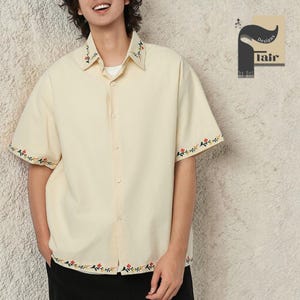 Op de afbeelding: Crèmekleurig shirt met korte mouwen en bloemenborduurwerk op de kraag en mouwranden. Het shirt heeft een knoopsluiting en een relaxte pasvorm. Het shirt maakt deel uit van de "Tair Designs by Bel" collectie.
