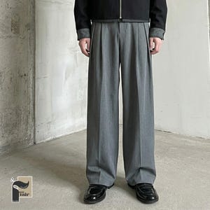 May include: A person wearing a black jacket and gray pleated trousers. The trousers are wide-legged and paired with black loafers. The outfit is set against a neutral background.
