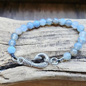 May include: A beaded bracelet featuring light blue and gray round beads. The bracelet is secured with a silver-colored hook clasp that reads "Hook". The bracelet is displayed on a piece of weathered wood.