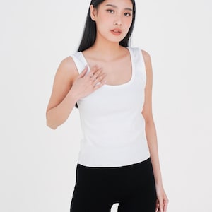 May include: A white tank top with a scoop neckline and wide straps. The top is paired with black leggings. The image is set against a plain white background, highlighting the simple design of the clothing.