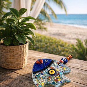 Pu&ograve; includere: Un pesce decorativo in vetro con un design a mosaico, con pezzi di vetro blu, giallo, rosso e trasparente. Il pesce ha una testa blu e un occhio giallo. Una piccola pianta in un cestino di vimini &egrave; nelle vicinanze.