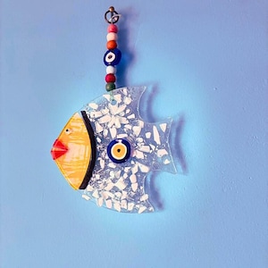 A vibrant handmade blue glass fish ornament featuring sunny yellow cheeks and a large evil eye bead for protection. This unique fused glass suncatcher reflects bright colors when hung in a window, making it a cheerful piece of handmade art for any home.