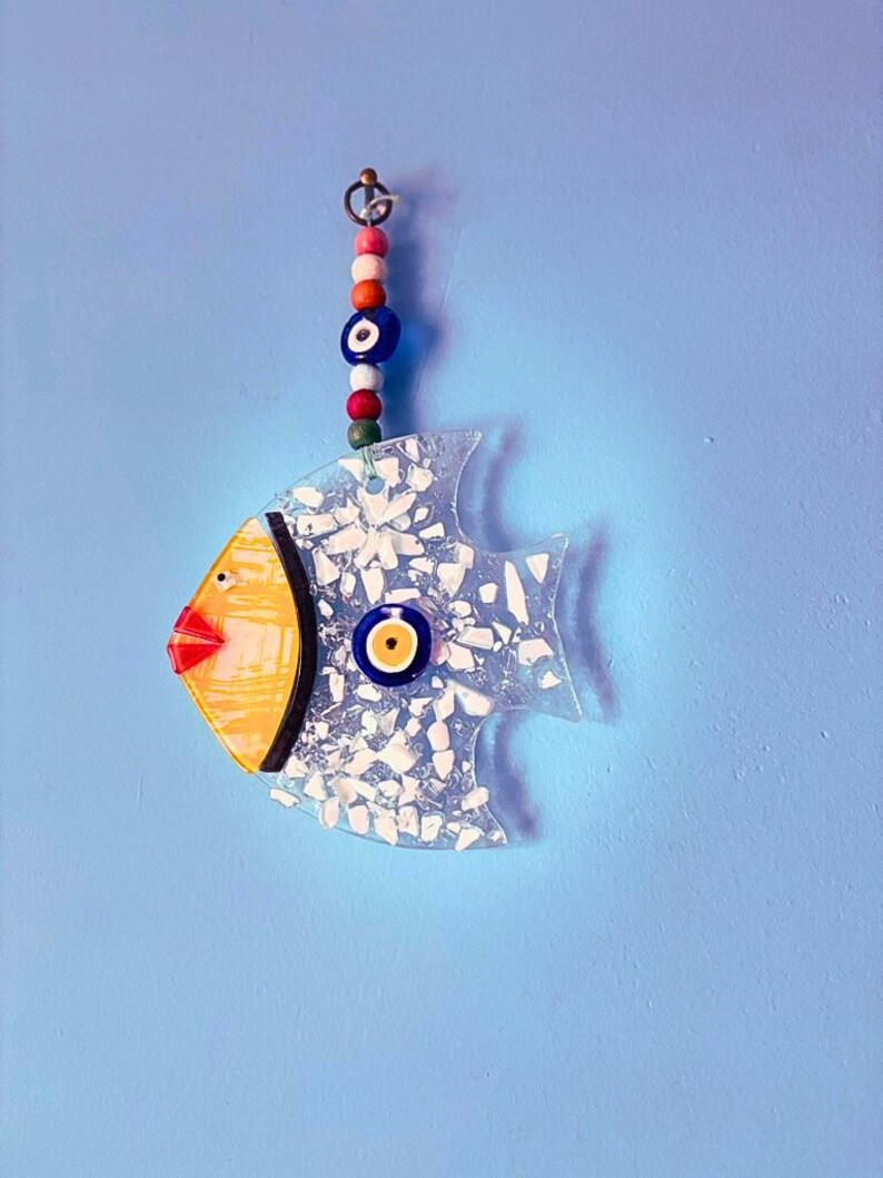 A vibrant handmade blue glass fish ornament featuring sunny yellow cheeks and a large evil eye bead for protection. This unique fused glass suncatcher reflects bright colors when hung in a window, making it a cheerful piece of handmade art for any home.