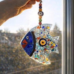 A colorful handmade glass fish suncatcher held against the sun. This vibrant fused glass wall hanging features blue, yellow, and red patterns that create beautiful light reflections for window decor.