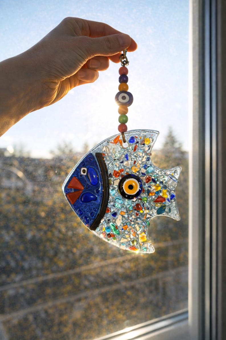 A colorful handmade glass fish suncatcher held against the sun. This vibrant fused glass wall hanging features blue, yellow, and red patterns that create beautiful light reflections for window decor.