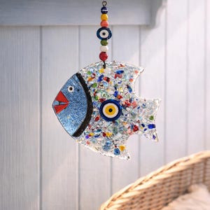 Pu&ograve; includere: Un ornamento decorativo a forma di pesce con testa blu, bocca rossa e corpo pieno di pezzi di vetro colorati. Presenta un ciondolo a forma di occhio e viene sospeso da una corda di perline. Misura circa 15 cm di lunghezza.