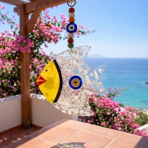Pu&ograve; includere: Un carillon a vento decorativo a forma di pesce in vetro, con testa gialla e corpo trasparente. Ha un occhio blu ed &egrave; sospeso a una corda di perline colorate. Lo sfondo mostra una vista sul mare e fiori rosa.