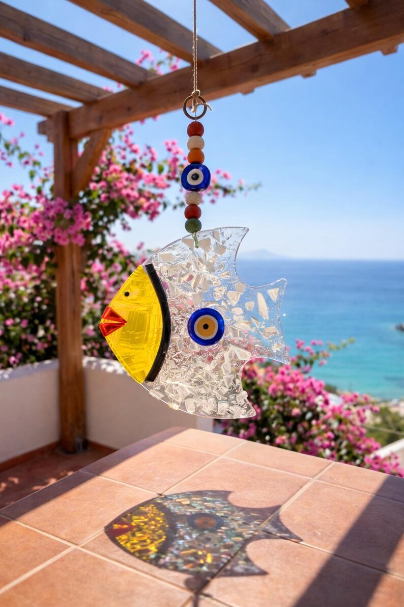 Pu&ograve; includere: Un carillon a vento decorativo a forma di pesce in vetro, con testa gialla e corpo trasparente. Ha un occhio blu ed &egrave; sospeso a una corda di perline colorate. Lo sfondo mostra una vista sul mare e fiori rosa.