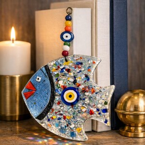 Pu&ograve; includere: Un pesce decorativo in vetro con un design a mosaico, con accenti blu, rossi e gialli. Il pesce ha una testa blu e un grande occhio al centro. Una catena di perline colorate &egrave; appesa in alto. Il pesce &egrave; lungo circa 15 cm.