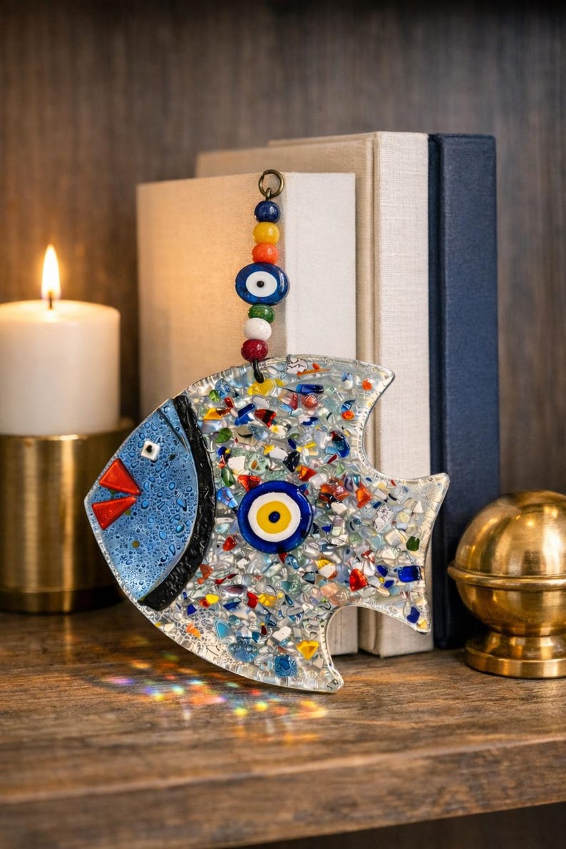 Pu&ograve; includere: Un pesce decorativo in vetro con un design a mosaico, con accenti blu, rossi e gialli. Il pesce ha una testa blu e un grande occhio al centro. Una catena di perline colorate &egrave; appesa in alto. Il pesce &egrave; lungo circa 15 cm.