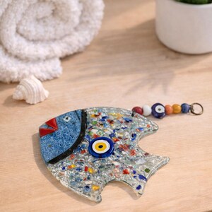 Pu&ograve; includere: Un pesce decorativo in vetro con testa blu e corpo a mosaico. Il pesce ha un occhio blu e giallo e una coda di perline. &Egrave; appoggiato su una superficie di legno, con un asciugamano bianco arrotolato e una pianta in vaso sullo sfondo.