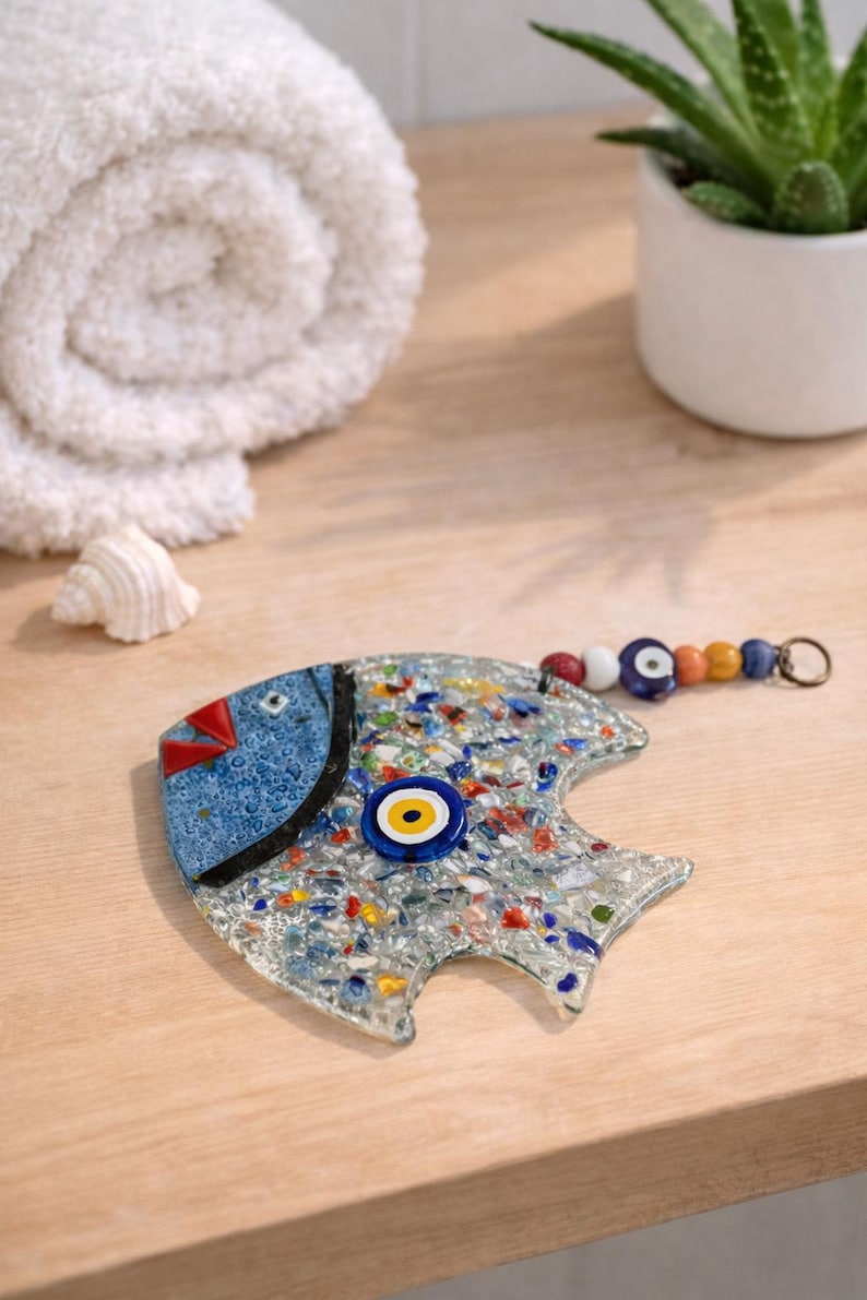 Pu&ograve; includere: Un pesce decorativo in vetro con testa blu e corpo a mosaico. Il pesce ha un occhio blu e giallo e una coda di perline. &Egrave; appoggiato su una superficie di legno, con un asciugamano bianco arrotolato e una pianta in vaso sullo sfondo.