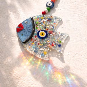Pu&ograve; includere: Un ornamento decorativo a forma di pesce con testa blu, labbra rosse e corpo pieno di pezzi di vetro colorati. Presenta un ciondolo a forma di occhio e una catena di perline per appenderlo. L'ornamento &egrave; lungo circa 13 cm.