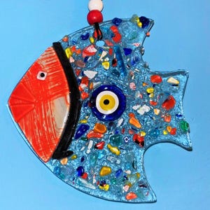 A cute handmade blue glass fish suncatcher with rosy red cheeks and a traditional evil eye bead on its body. This charming fused glass wall art features colorful patterns and vibrant details, making it a perfect nursery decor or a cheerful housewarming gift.
