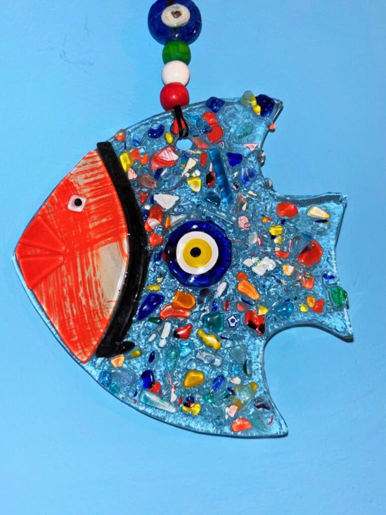 A cute handmade blue glass fish suncatcher with rosy red cheeks and a traditional evil eye bead on its body. This charming fused glass wall art features colorful patterns and vibrant details, making it a perfect nursery decor or a cheerful housewarming gift.