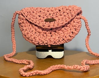 May include: A handmade, light pink crochet crossbody bag with a flap closure. The bag features a braided strap and a small, rectangular tag that reads "HANDMADE". The bag is displayed on a wooden surface.