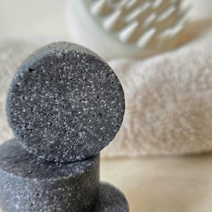 May include: Three dark gray, round soap bars stacked on top of each other. The soap bars have a speckled texture. A white, textured scalp massager and a beige towel are in the background.