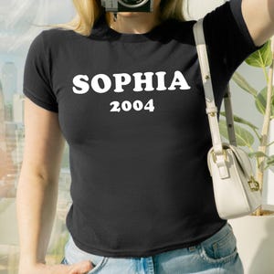 May include: Black t-shirt with the name "SOPHIA" and the year "2004" printed in white. The person is wearing a white cap and holding a camera. A white purse is hanging from the shoulder. The text "Send your text. We send preview before printing" is in a white box.