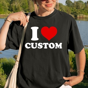 Può includere: Una t-shirt nera a maniche corte con il testo bianco I, un simbolo di cuore rosso e il testo bianco CUSTOM stampato sul davanti. La maglietta presenta un girocollo e una vestibilità rilassata, abbinata a pantaloncini bianchi e una borsa a tracolla beige.
