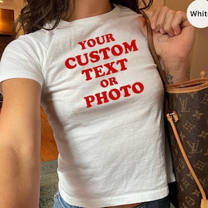 May include: White t-shirt with the text "YOUR CUSTOM TEXT OR PHOTO" in red. The shirt is worn with blue jeans. A brown handbag with a monogram pattern is visible.