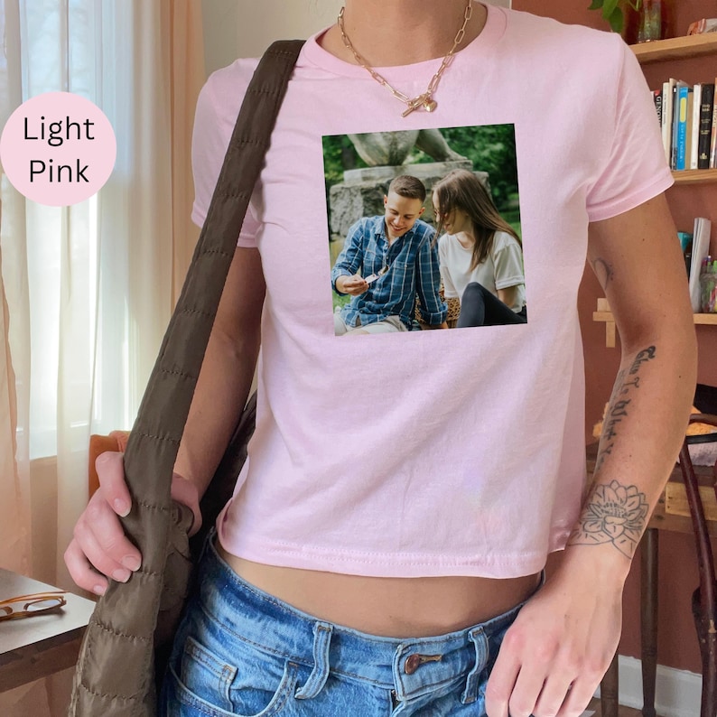 May include: Light pink crop top t-shirt with a square image of two people. The image shows a man in a blue plaid shirt and a woman in a white shirt. The t-shirt is worn with blue jeans.