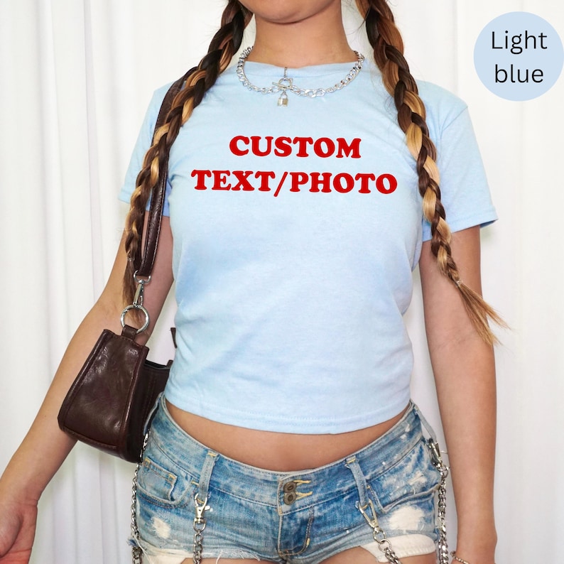 May include: Light blue crop top with the text "CUSTOM TEXT/PHOTO" in red. The shirt is short-sleeved and has a round neckline. A light blue circle with the words "Light blue" is in the upper right corner.