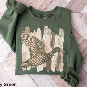 May include: A military green long-sleeve sweatshirt featuring a central graphic of a flying duck. The duck's body is decorated with a mix of checkered, leopard print, and polka dot patterns. Behind the duck is a beige and brown brushstroke design.