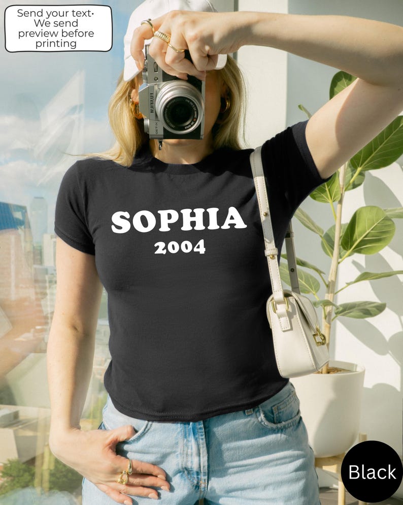 May include: Black t-shirt with the name "SOPHIA" and the year "2004" printed in white. The person is wearing a white cap and holding a camera. A white purse is hanging from the shoulder. The text "Send your text. We send preview before printing" is in a white box.