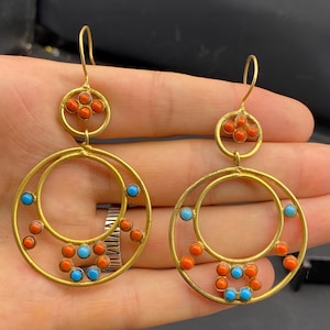 Könnte beinhalten: Ein Paar goldfarbene Ohrringe mit kreisförmigem Design. Jeder Ohrring hat zwei konzentrische Kreise, die mit orangefarbenen und türkisfarbenen Perlen verziert sind. Die Ohrringe werden von einem Haken gehalten.