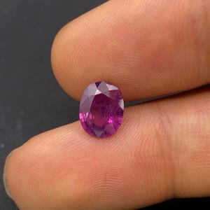 Puede incluir: Una piedra preciosa facetada de forma ovalada con un tono magenta y púrpura intenso. La piedra presenta una superficie pulida con múltiples cortes geométricos que reflejan la luz, mostrando una calidad translúcida y un color vibrante.
