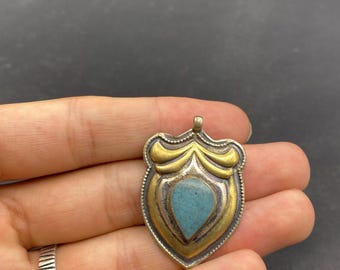 May include: A shield-shaped pendant with a turquoise stone center. The pendant has a silver and gold-tone metal design. The turquoise stone is teardrop-shaped. The pendant is held in a hand.