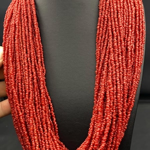 May include: A striking red beaded necklace presented on a black display stand. The necklace features multiple strands of small, round beads, giving a full, textured look. The beads are a consistent, bright red, and the necklace is arranged in a U-shape.