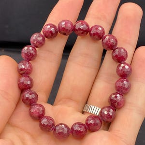 May include: A bracelet made of deep red, faceted gemstones. The round beads are strung together to form a complete circle. The bracelet is held in a hand, highlighting the rich colour and texture of the stones.