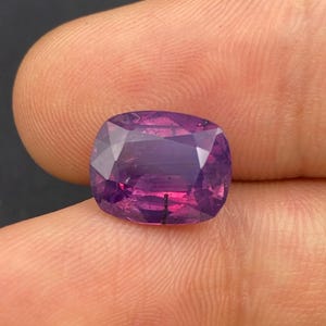 Puede incluir: Una piedra preciosa de corte cojín con un tono púrpura y magenta intenso, con superficies facetadas que reflejan la luz. La piedra muestra una claridad transparente con inclusiones internas visibles y una marca lineal oscura central.