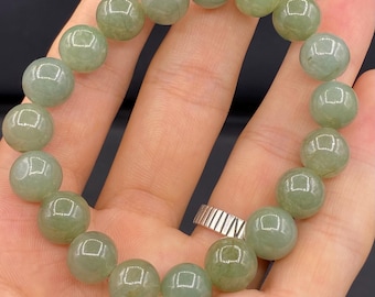 May include: A jade bracelet featuring round, translucent, light green beads. The beads are strung together to form a circular bracelet. The bracelet is displayed on a hand, with a silver ring visible.
