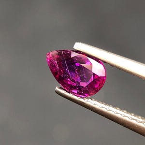 Puede incluir: Una gema facetada en forma de pera, sujeta con pinzas plateadas. La gema es de color magenta intenso, con facetas internas que reflejan la luz. El fondo es gris oscuro.