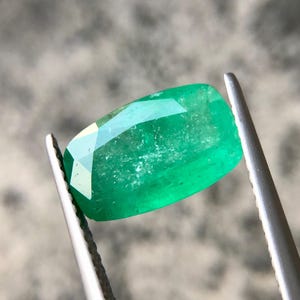 以下が含まれることがあります： ピンセットで保持された鮮やかな緑色のエメラルドの宝石。エメラルドは長方形で、光を反射するファセットエッジがあります。宝石の色は豊かで深い緑色で、その自然な美しさを際立たせています。クローズアップビューは、宝石の透明度と輝きを強調しています。