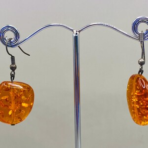 Può includere: Un paio di orecchini in ambra con ganci d'argento. L'ambra è di un colore arancione caldo e ha un aspetto naturale e organico.