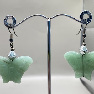 以下が含まれることがあります： 銀色のイヤリングスタンドに掛けられた、銀色のアクセントが付いた緑色の蝶の形をしたイヤリングのペア