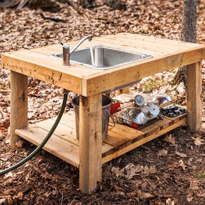 Könnte beinhalten: Ein hölzernes Spielwaschbecken für den Außenbereich mit einem Edelstahlbecken und Wasserhahn. Das Waschbecken steht auf einem Holztisch mit einem unteren Regal. Ein grüner Schlauch und ein Metalleimer sind angeschlossen. Verschiedene Küchenartikel befinden sich auf dem Regal.